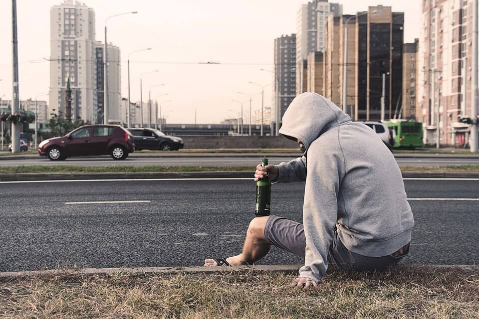 Man in hoodie drinking on roadside in urban area, symbolizing street alcohol abuse